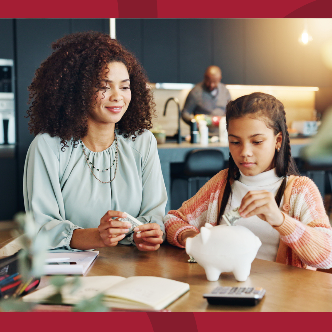 A mother and daughter are sitting at the kitchen table.  The daughter is putting money into a piggy bank.